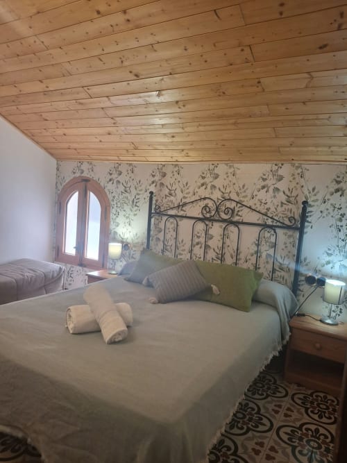 Cozy bedroom at Refugio la Presa with wrought-iron bed, wooden ceiling, botanical wallpaper, and arched window with landmark