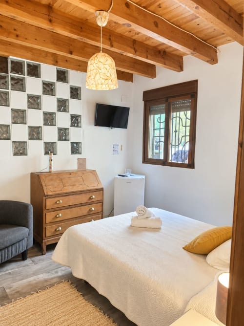 Cozy bedroom at Refugio la Presa, Chulilla with wooden beams, white bed, wooden dresser, TV, and decorative pendant light
