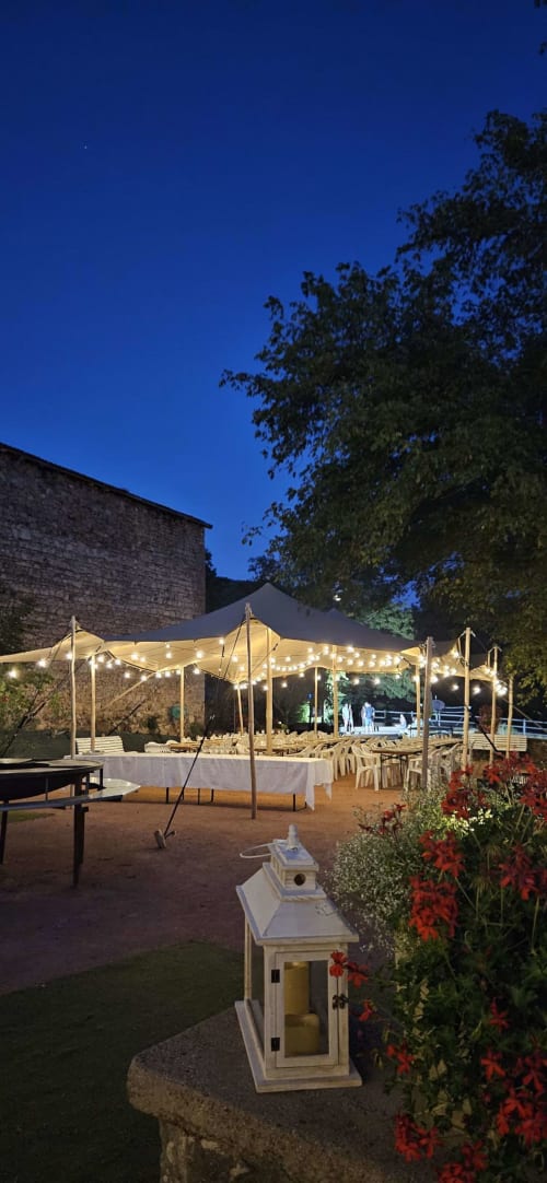 Gîtes Les Muriers : soirée en plein air avec tente illuminée, places assises, fleurs rouges et bâtiments en pierre à Bourgvilain