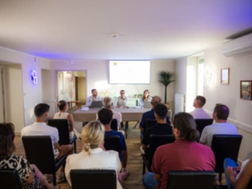 Salle de conférence aux Gites Les Muriers avec participants assis face à un panel d'orateurs à une table sous un écran de projection.