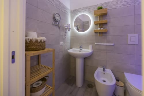 Moderno bagno presso l'Aurum Nolana Suites a Pompei con specchio illuminato, lavabo su colonna, bidet, scaffalature in legno e piastrelle