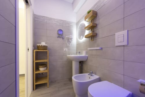 Bagno moderno in Aurum Nolana Suites Pompeii con lavabo bianco, bidet, wc, ripiani in legno e illuminazione con tonalità viola