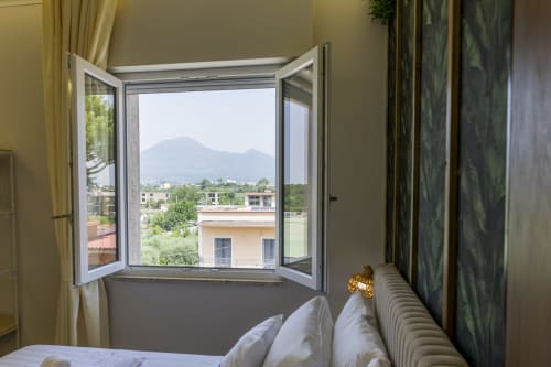 Finestre aperte in camera d'hotel a Pompei con vista sul Vesuvio, letto con cuscini, tende e balcone affacciato sulla città