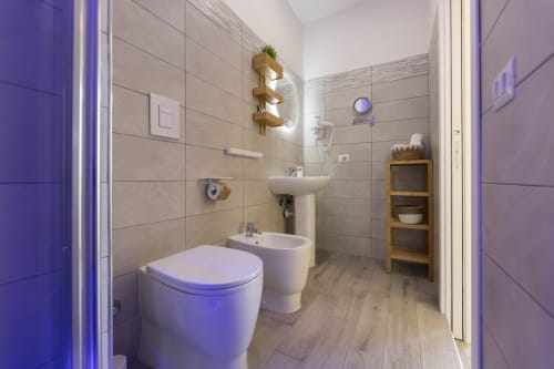 Bagno moderno nelle Aurum Nolana Suites a Pompei con wc bianco, bidet, lavabo su piedistallo, scaffali in legno e illuminazione viola