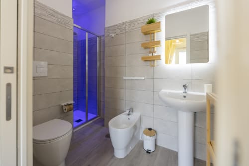 Bagno moderno in Aurum Nolana Suites Pompei con doccia illuminata blu, lavabo bianco, bidet, specchio retroilluminato e scaffali in legno