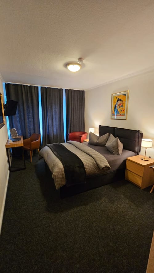 Modernes Hotelzimmer im The Soho Hotel Düsseldorf mit Queensize-Bett, dunklen Vorhängen, Holzmöbeln und zeitgenössischer Einrichtung.