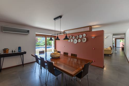 Sala de jantar moderna na quinta Trindade com mesa de madeira, cadeiras pretas, parede em tons de terracota, lustres pendentes e vista para a piscina