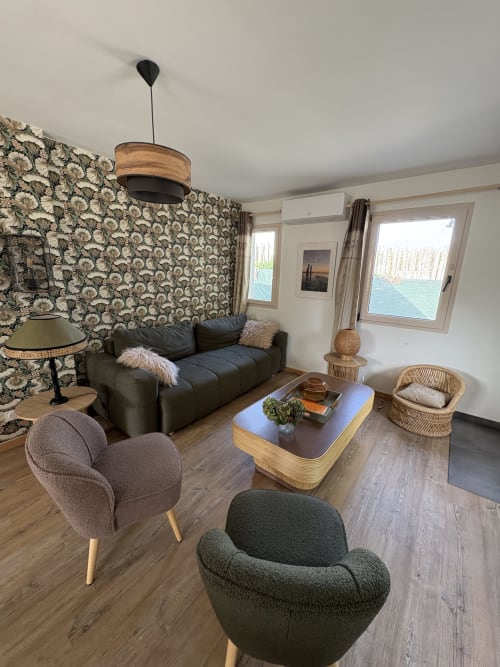 Salon douillet du Village Du Moulin avec papier peint fleuri, canapé en cuir foncé, fauteuils arrondis, table basse en bois