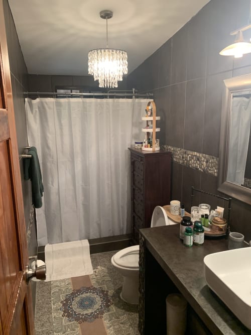 Modern bathroom with dark tile walls, crystal pendant light, dark vanity, white sink, toilet, and shower with gray curtain at