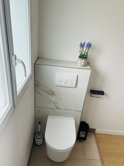 Salle de bains moderne avec toilettes blanches, vasque en marbre, fleurs violettes dans un pot tressé et fenêtre à La villa Cassinne