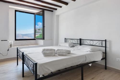 Bright bedroom with black metal bed, white linens, and open window overlooking Caccamo mountains at Sikania Agri Relais