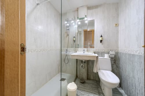 Baño moderno con ducha abierta, lavabo blanco, inodoro y cenefa de azulejos decorativos en el Hostal El Molino de Collado Villalba