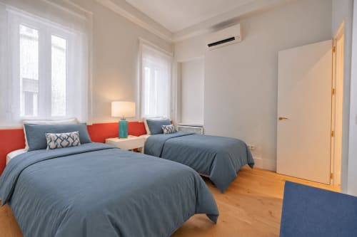 Dormitorio moderno en Madrid con dos camas, ropa de cama azul, cabeceros rojos, suelo de madera y vistas a la ciudad desde grandes ventanales