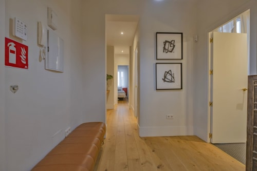 Moderno pasillo de apartamento en Madrid con suelos de madera, banco de cuero, cuadros enmarcados y puertas blancas hacia las zonas de estar