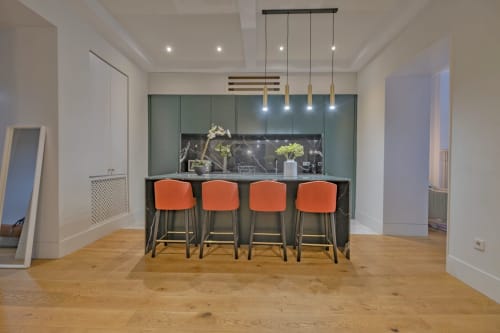Moderna cocina de apartamento madrileño con isla, taburetes naranjas, iluminación colgante y azulejos verdes de salpicadero