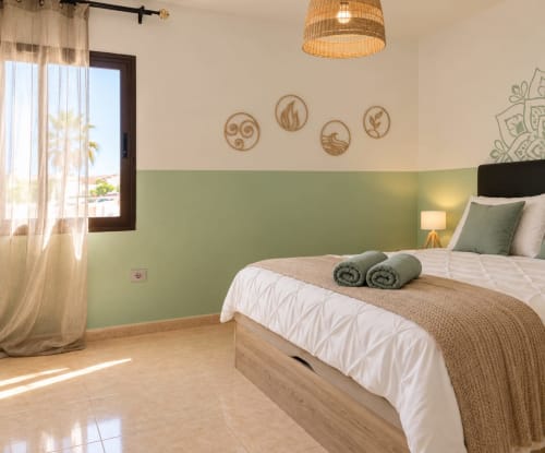 Helles Schlafzimmer bei Sara & Gino Fuerteventura mit Salbeigrüner Akzentwand, Holzbett, gewebter Deckenleuchte und Gartenblick