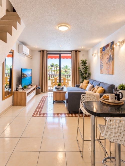 Modern living room in Tenerife apartment with gray sofa, wooden furniture, palm tree views from balcony, TV, and beige tile