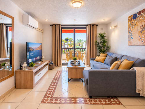 Bright modern apartment living room with gray sofa, TV, wooden furniture, and balcony view of palm trees in Tenerife