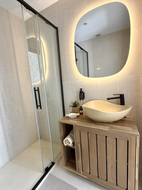 Modern bathroom with illuminated rounded mirror, white vessel sink, wooden vanity, and glass shower enclosure at HomyMarbella