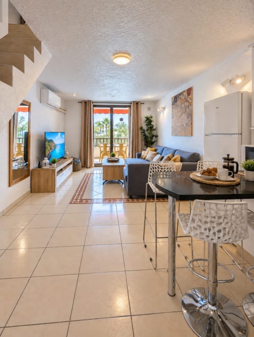 Modern open-plan apartment living area with kitchen, dining counter, sofa, TV, and balcony overlooking palm trees in Tenerife