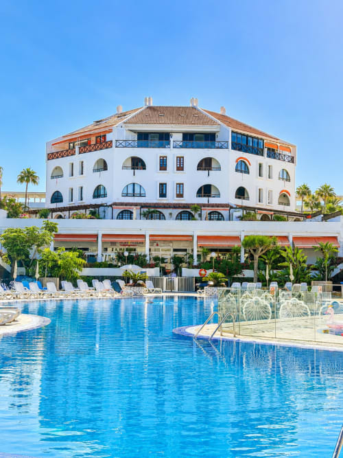 Beachfront hotel in Tenerife with white architecture, terracotta roof, blue pool, and palm trees under clear sky
