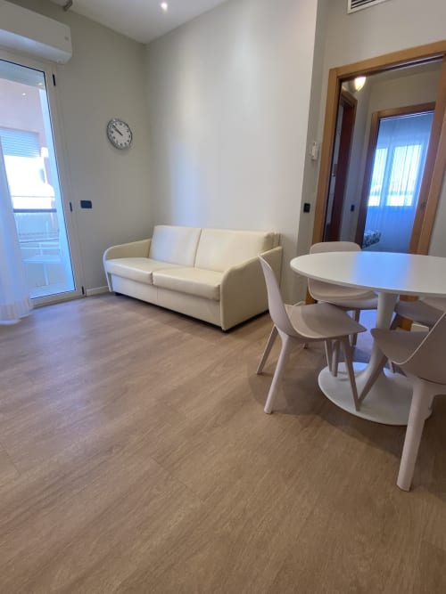 Soggiorno moderno nell'appartamento Comfort dell'hotel Rimini con divano color crema, tavolo rotondo bianco e pavimenti in legno