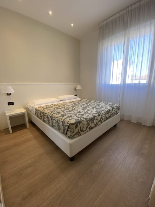 Camera moderna di Residence Aida Rimini con letto bianco, piumone a motivi, pavimenti in legno e tende leggere luminose