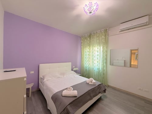Camera da letto dell'appartamento Deledda a Firenze con parete accent viola, letto bianco, tende verdi e servizi moderni