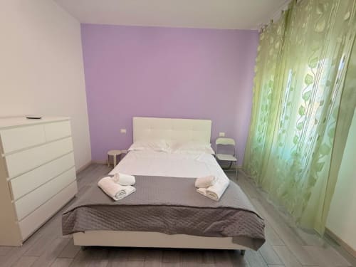Camera moderna in un appartamento fiorentino con letto bianco, parete d'accento viola, tende verdi, cassettiera e pavimento piastrellato