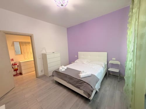 Camera da letto moderna nell'appartamento Deledda a Firenze con letto bianco, parete accent viola, tende verdi e vista sulla città