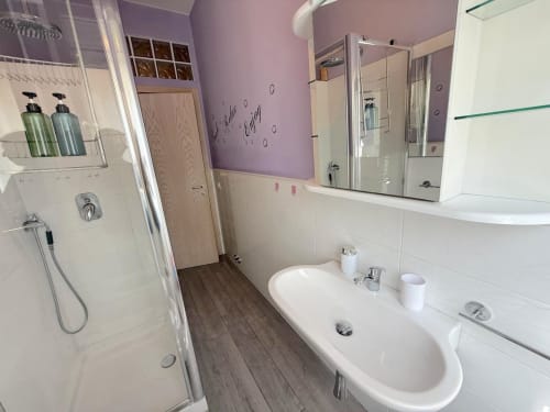 Bagno moderno dell'appartamento Deledda a Scandicci, con pareti accent viola, doppio lavabo, doccia e piastrelle bianche