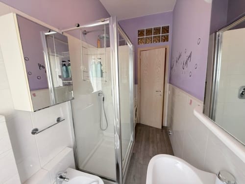 Bagno moderno nell'appartamento Deledda a Scandicci con muri viola, cabina doccia in vetro, WC e piastrelle bianche