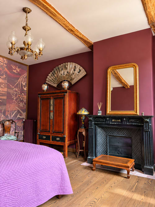 Chambre élégante à la Maison Crone avec murs bordeaux, poutres en bois, miroir orné, cheminée et lit violet