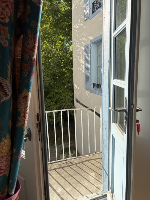 Portes-fenêtres ouvertes de la Chambre Eva donnant sur un balcon ensoleillé aux balustrades blanches surplombant les jardins verdoyants de la Maison