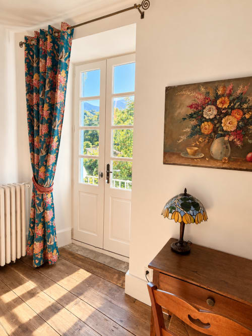 Chambre ensoleillée de la Maison Crone avec rideaux à motifs floraux bleus, portes-fenêtres donnant sur le jardin, lampadaire vintage et tableau nature morte