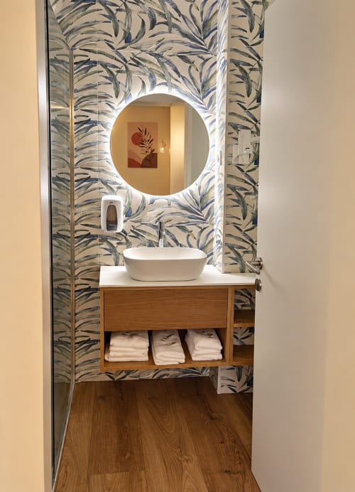Banheiro moderno com cuba cilíndrica branca, espelho redondo iluminado, armário de madeira e papel de parede botânico azul