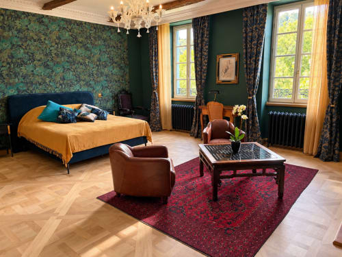 Chambre Émeraude de la Maison Crone avec papier peint émeraude, literie dorée, fauteuils en cuir et vue sur le jardin