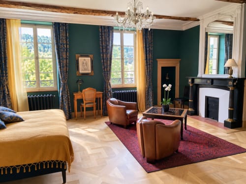 Chambre Émeraude à Maison Crone : élégante chambre aux murs verts, cheminée, sièges en cuir, lustre, vue sur le jardin