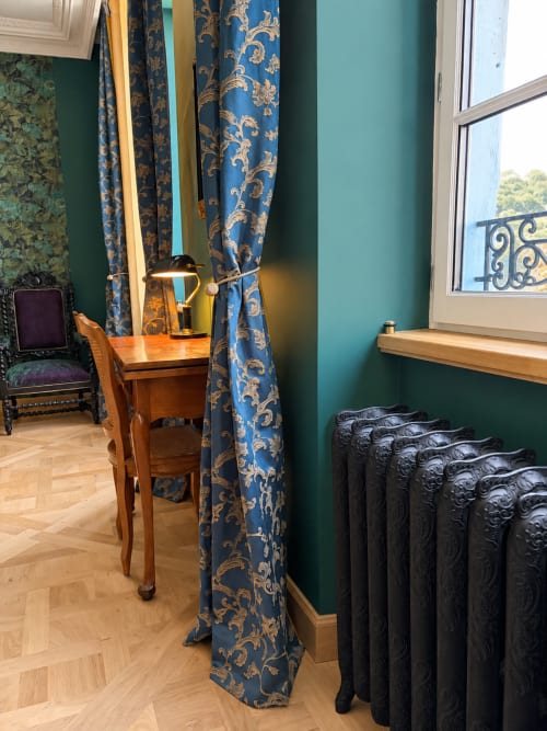 Chambre émeraude à la Maison Crone avec rideaux damassés bleus, bureau en bois, radiateur ouvragé et vue sur le jardin