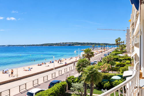 Vue du littoral de Cannes : plage de sable fin, eaux cristallines de la Méditerranée, palmiers et élégant bâtiment hôtelier blanc