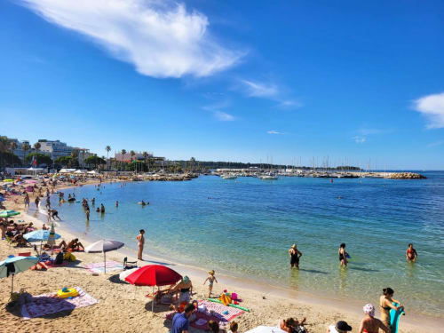 Plage animée de Cannes avec eaux turquoise, baigneurs, parasols et voiliers dans le port sous ciel bleu