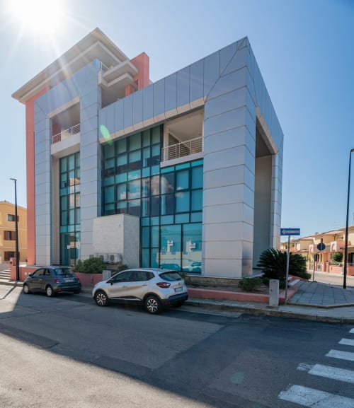 Modern Short Let's Olbia building with blue glass facade, red accents, and parked cars on street in Olbia, Italy