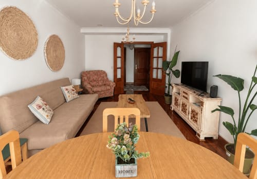 Apartamento Amieiro en Baiona: amplio salón-comedor con mesa de madera, sofá beige, TV, lámpara de araña y decoración natural.