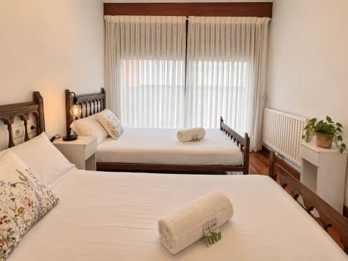 Dormitorio luminoso en apartamento Holibai, Baiona con dos camas de madera, ropa blanca y grandes ventanas con cortinas translúcidas