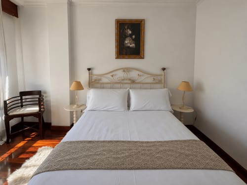 Dormitorio luminoso en apartamento de Baiona con sábanas blancas, cabecero metálico, lámparas de noche, suelo de madera y cuadros decorativos