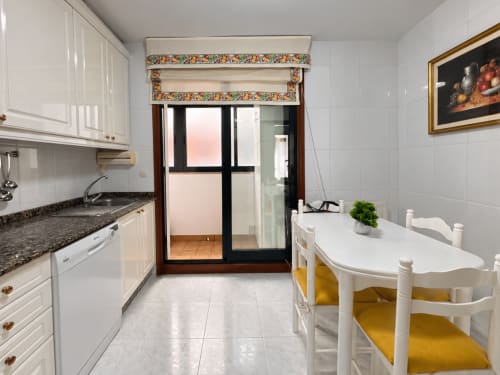 Luminosa sala de estar-comedor en apartamento de Baiona con armarios blancos, encimeras de granito, estor romano floral y mesa redonda