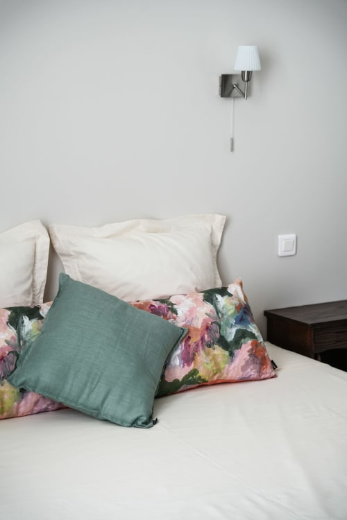 Le Saint Laurans bedroom with white bedding, green and floral pillows, wall lamp, and light switch in Aurignac