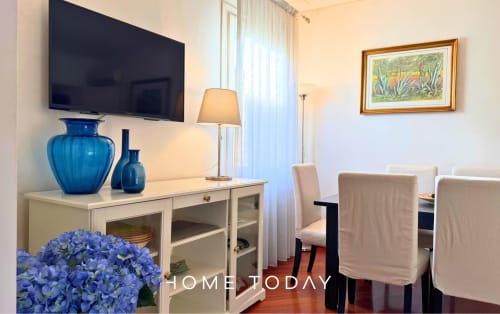 Modern apartment dining area in Padova with blue hydrangeas, white cabinetry, mounted TV, and neutral dining chairs