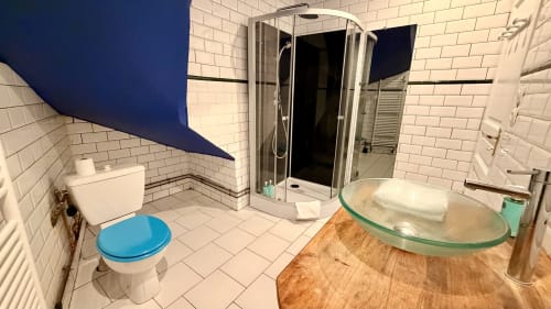 Salle de bain moderne avec abattant toilettes bleu, vasque en verre sur comptoir bois et douche ouverte à la villa Maison Agami