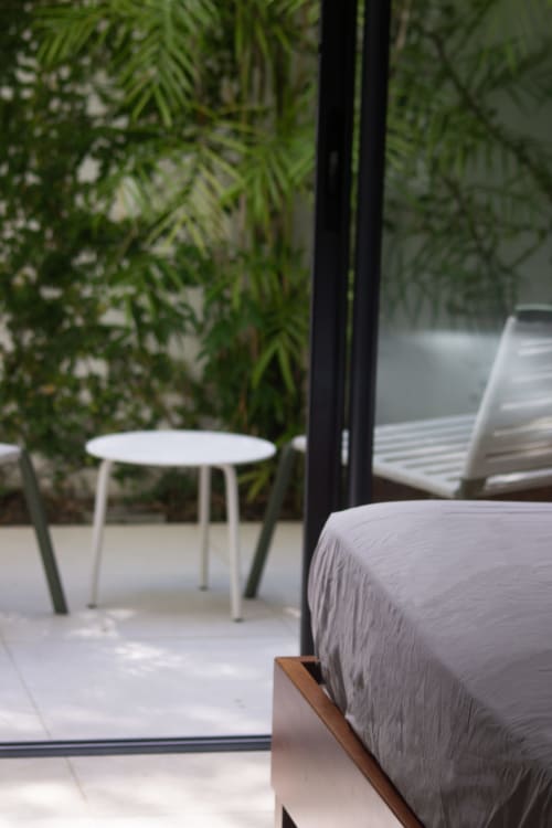 Chambre de villa bord de mer avec linge gris, table blanche, pergola noire et vue sur jardin tropical luxuriant à INHÉRENCE